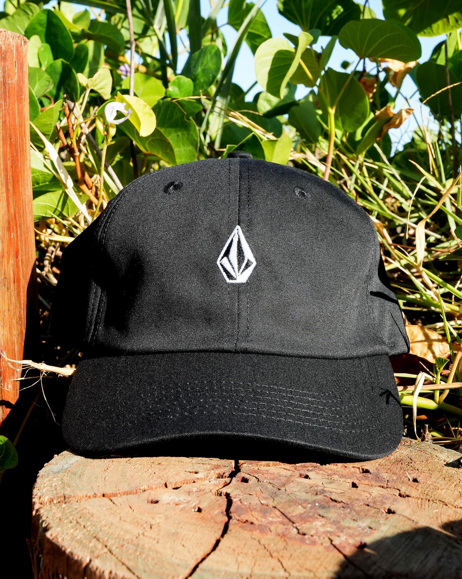 Imagem frontal do boné preto Volcom com logo branco, sobre base de madeira e folhas ao fundo, combinando estilo street com natureza.