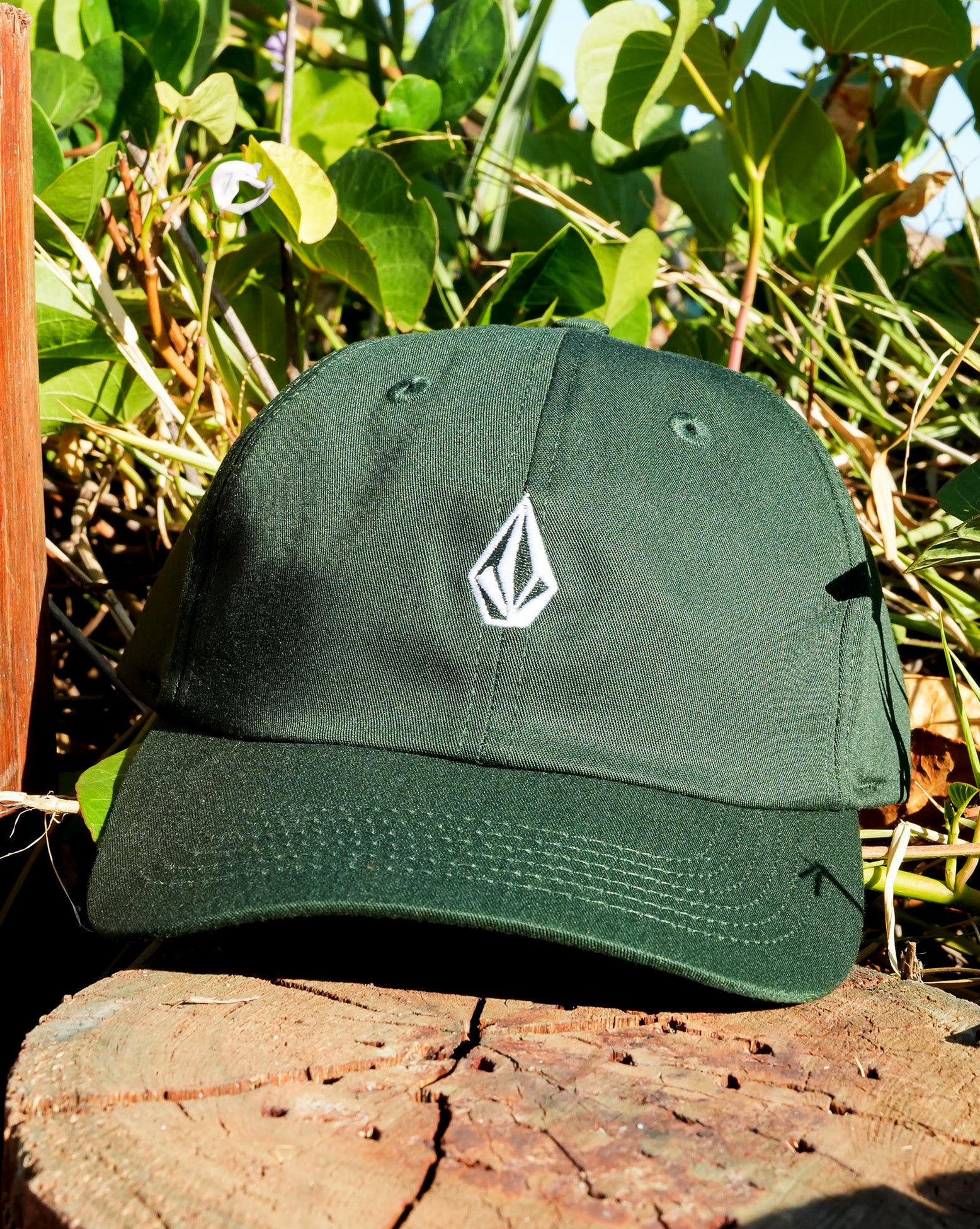 Imagem frontal do boné Volcom verde com logo bordado em branco, em meio à vegetação, destacando o estilo urbano e natural da peça.